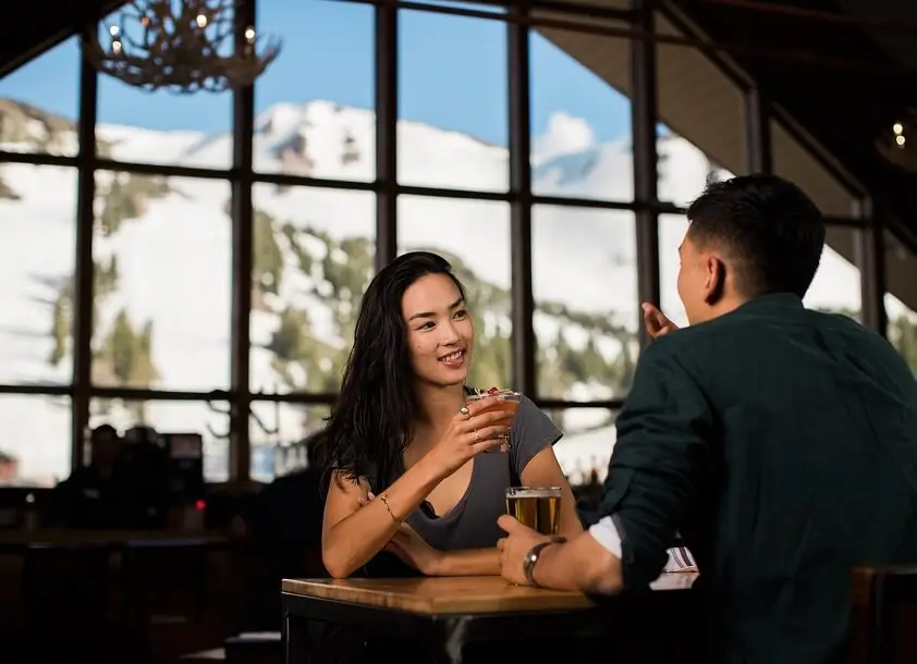 Image of a couple sips cocktails and beer in Main Lodge with Mammoth Mountain in the background